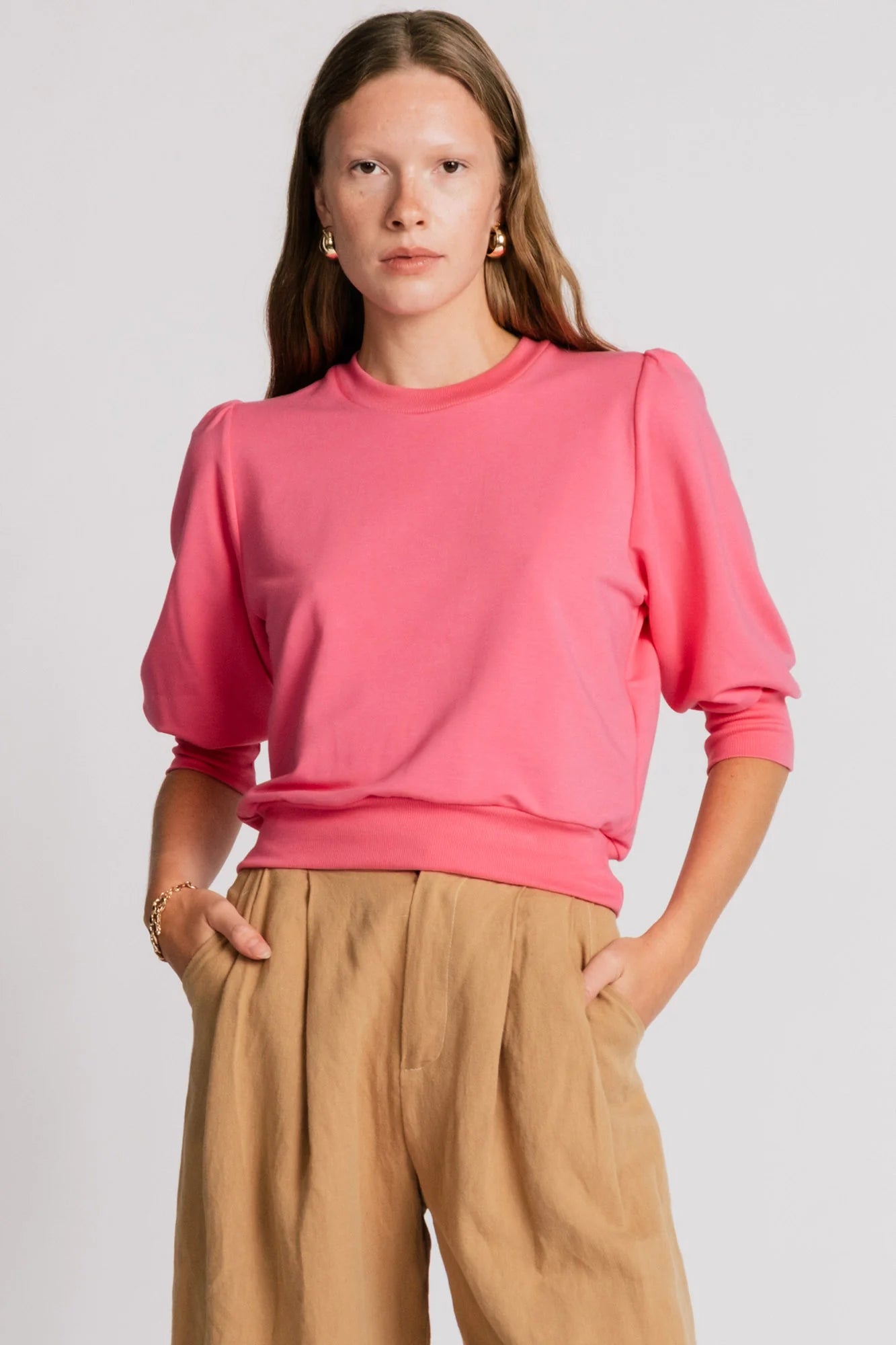 A woman wearing the Finch Sweatshirt by Allison Wonderland in Pink, a cropped sweatshirt with elbow-length puffed sleeves and ribbing at the neck and waistband. She is wearing it with beige pants and standing in front of a white background.