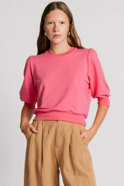 A woman wearing the Finch Sweatshirt by Allison Wonderland in Pink, a cropped sweatshirt with elbow-length puffed sleeves and ribbing at the neck and waistband. She is wearing it with beige pants and standing in front of a white background.