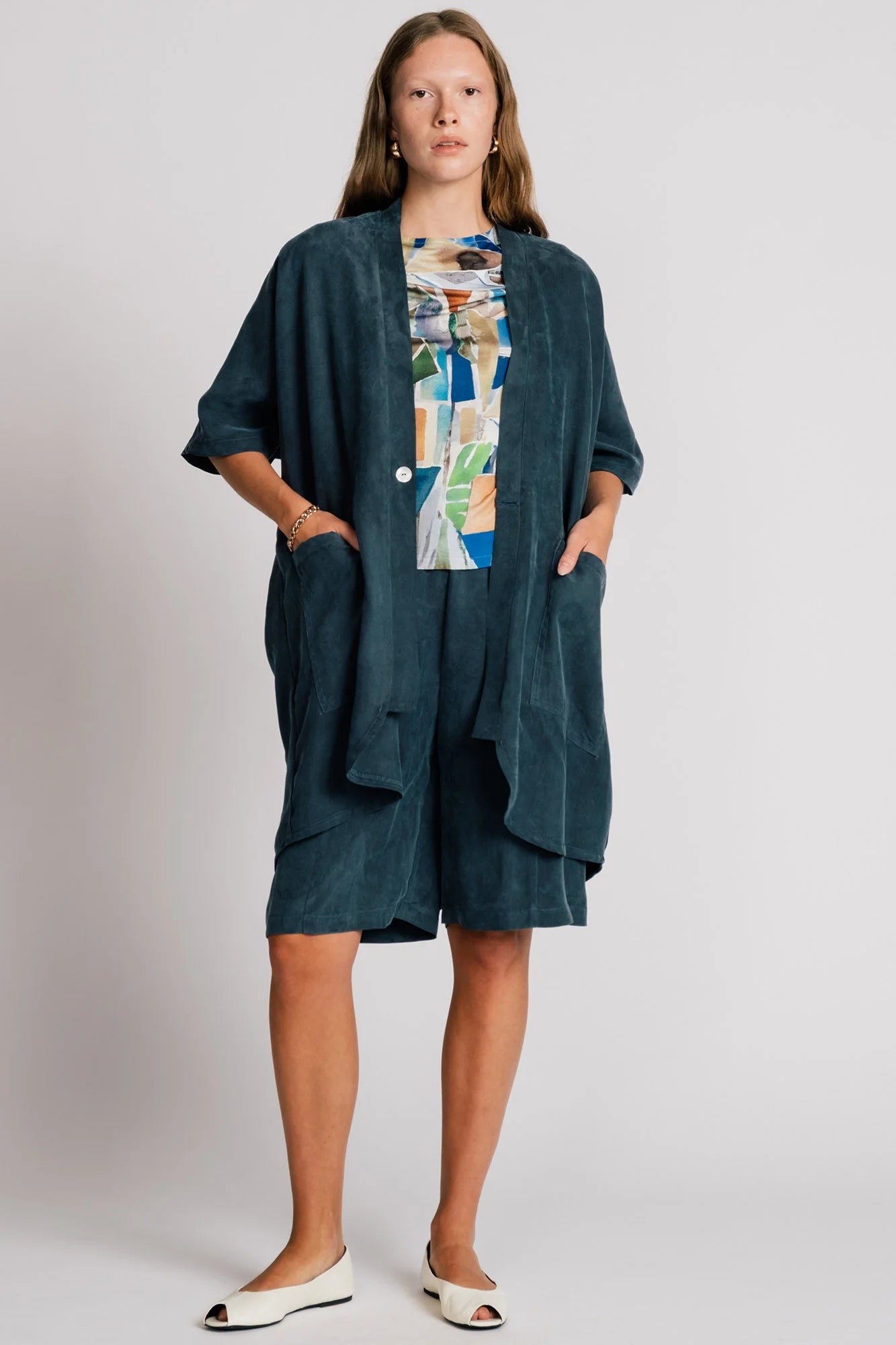 A woman wearing the Ibis Wrap from Allison Wonderland in Petrol blue, a robe-like jacket with elbow-length sleeves, a button closure and two large patch pockets. She is wearing it with a colourful top and matching shorts and standing in front of a white background. 