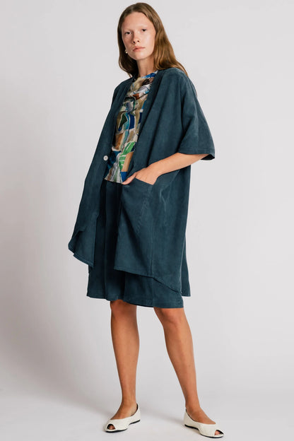 A woman wearing the Ibis Wrap from Allison Wonderland in Petrol blue, a robe-like jacket with elbow-length sleeves, a button closure and two large patch pockets. She is wearing it with a colourful top and matching shorts and standing in front of a white background. 