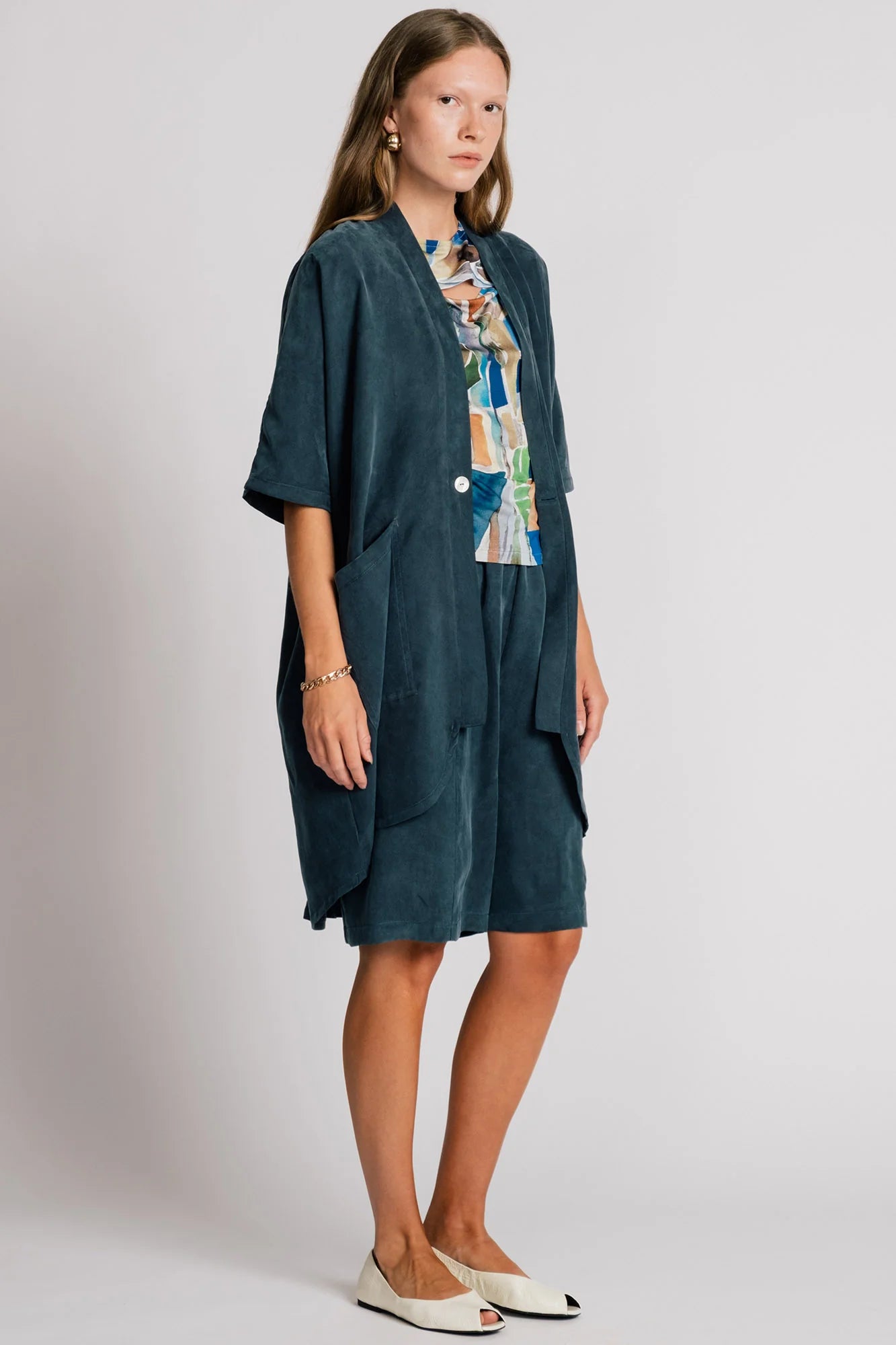 A woman wearing the Ibis Wrap from Allison Wonderland in Petrol blue, a robe-like jacket with elbow-length sleeves, a button closure and two large patch pockets. She is wearing it with a colourful top and matching shorts and standing in front of a white background. 
