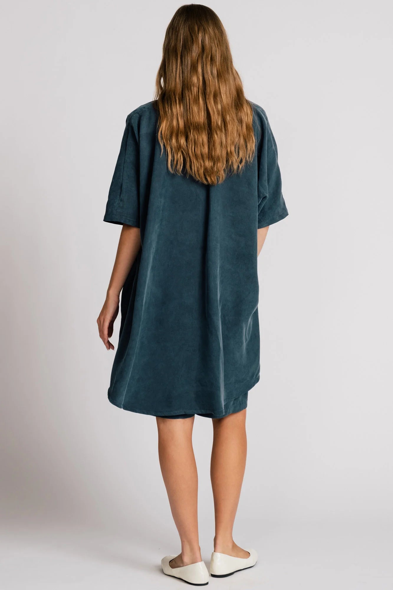 Back view of A woman wearing the Ibis Wrap from Allison Wonderland in Petrol blue, a robe-like jacket with elbow-length sleeves, a button closure and two large patch pockets. She is wearing it with a colourful top and matching shorts and standing in front of a white background. 