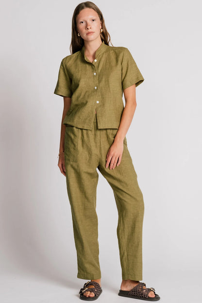 A woman wearing the Kite Blouse by Allison Wonderland in Avocado with subtle check print, a blouse with a button front and a banded collar. It has  slightly loose fit and short sleeves. She is wearing it with matching pants and standing in front of a white background. 