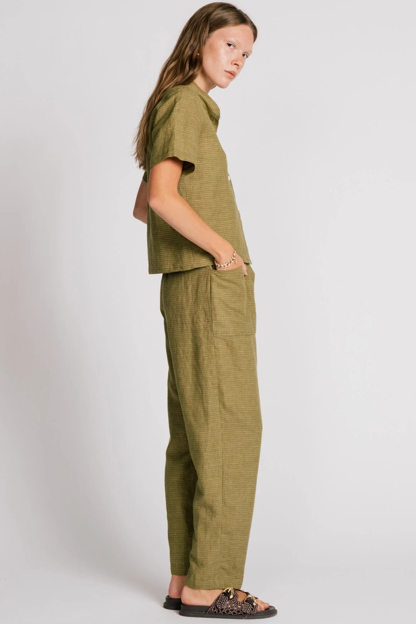 Side view of a woman wearing the Kite Blouse by Allison Wonderland in Avocado with subtle check print, a blouse with a button front and a banded collar. It has  slightly loose fit and short sleeves. She is wearing it with matching pants and standing in front of a white background. 