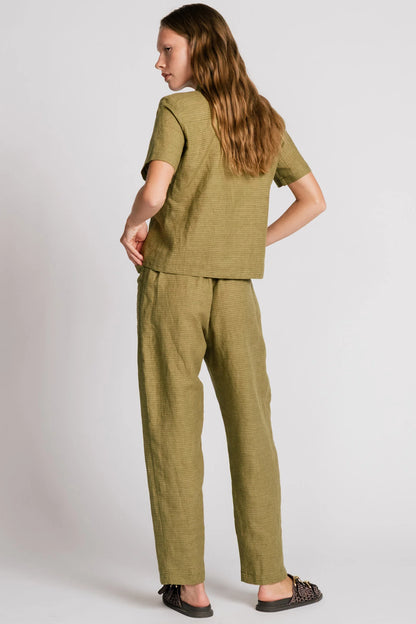 Back view of a woman wearing the Kite Blouse by Allison Wonderland in Avocado with subtle check print, a blouse with a button front and a banded collar. It has  slightly loose fit and short sleeves. She is wearing it with matching pants and standing in front of a white background. 