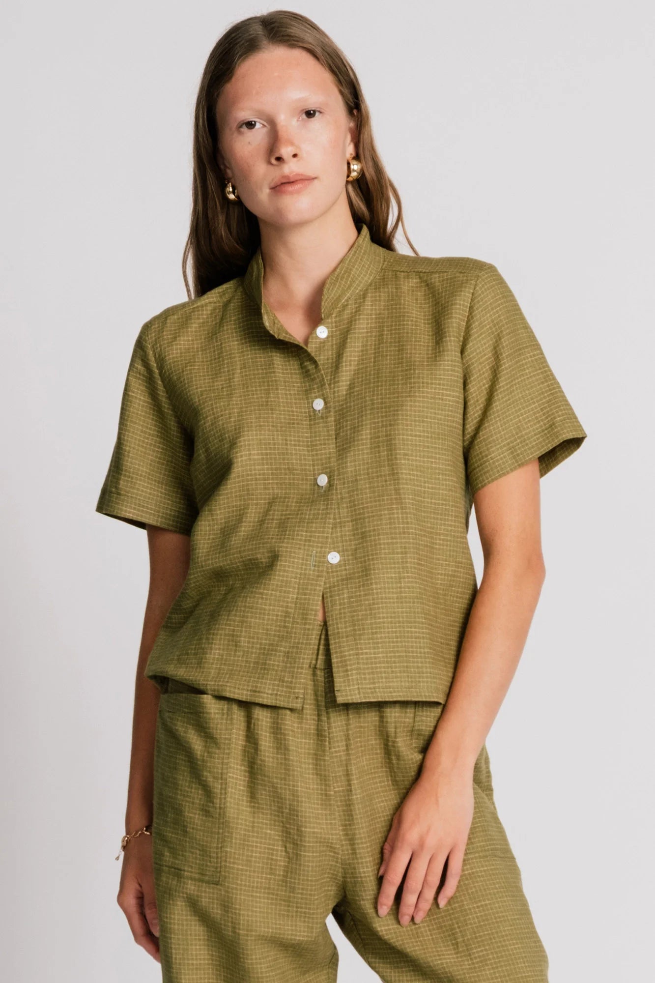 A woman wearing the Kite Blouse by Allison Wonderland in Avocado with subtle check print, a blouse with a button front and a banded collar. It has  slightly loose fit and short sleeves. She is wearing it with matching pants and standing in front of a white background. 