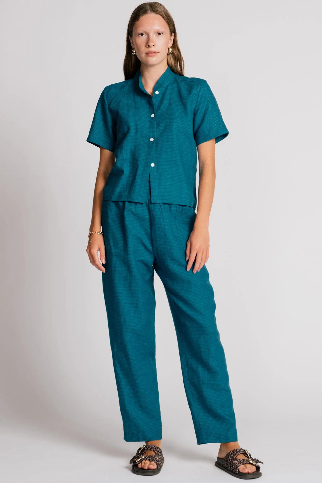 A woman wearing the Kite Blouse by Allison Wonderland in Teal with subtle check print, a blouse with a button front and a banded collar. It has  slightly loose fit and short sleeves. She is wearing it with matching pants and standing in front of a white background. 