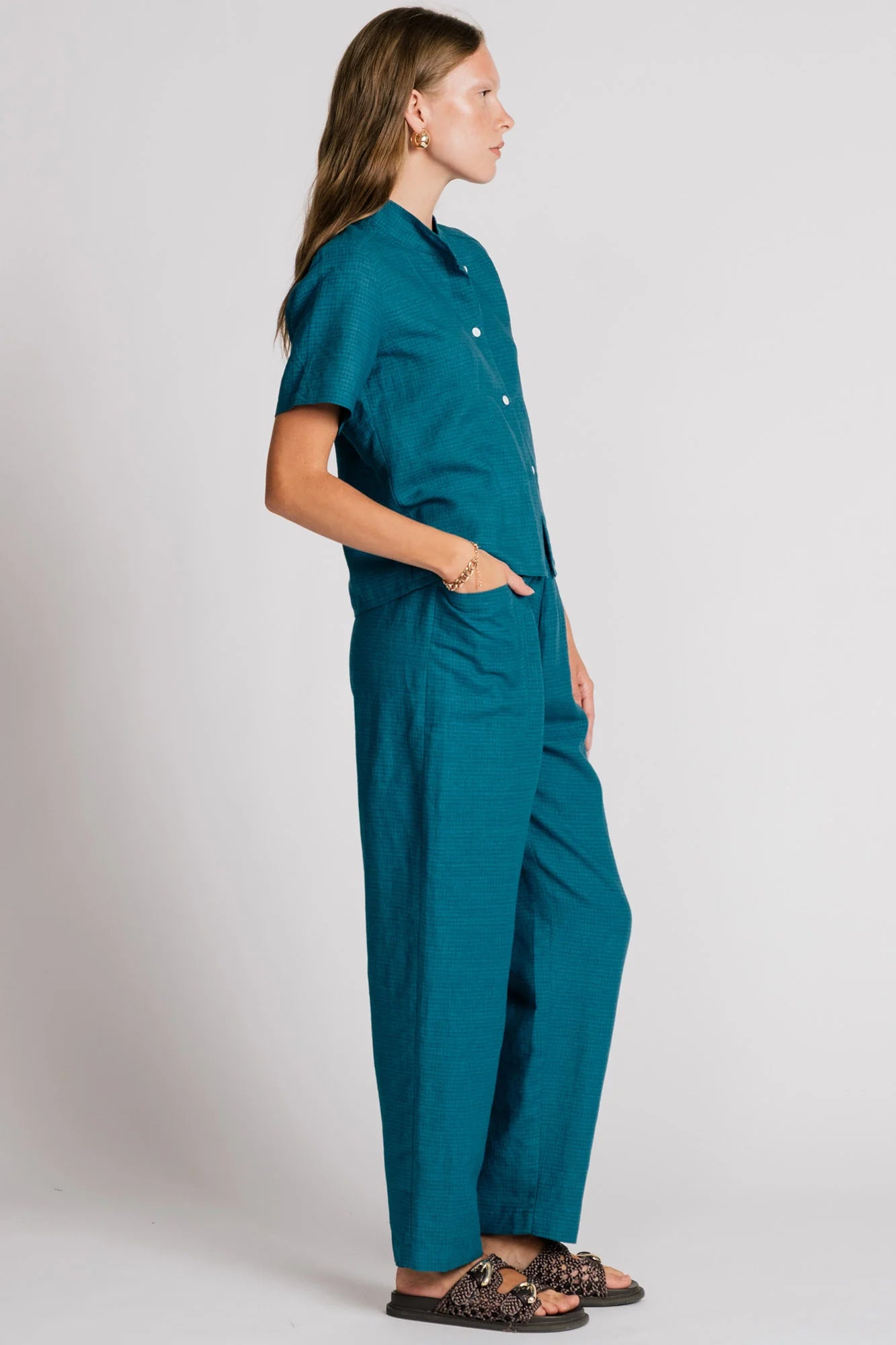 Side view of a woman wearing the Kite Blouse by Allison Wonderland in Teal with subtle check print, a blouse with a button front and a banded collar. It has  slightly loose fit and short sleeves. She is wearing it with matching pants and standing in front of a white background. 