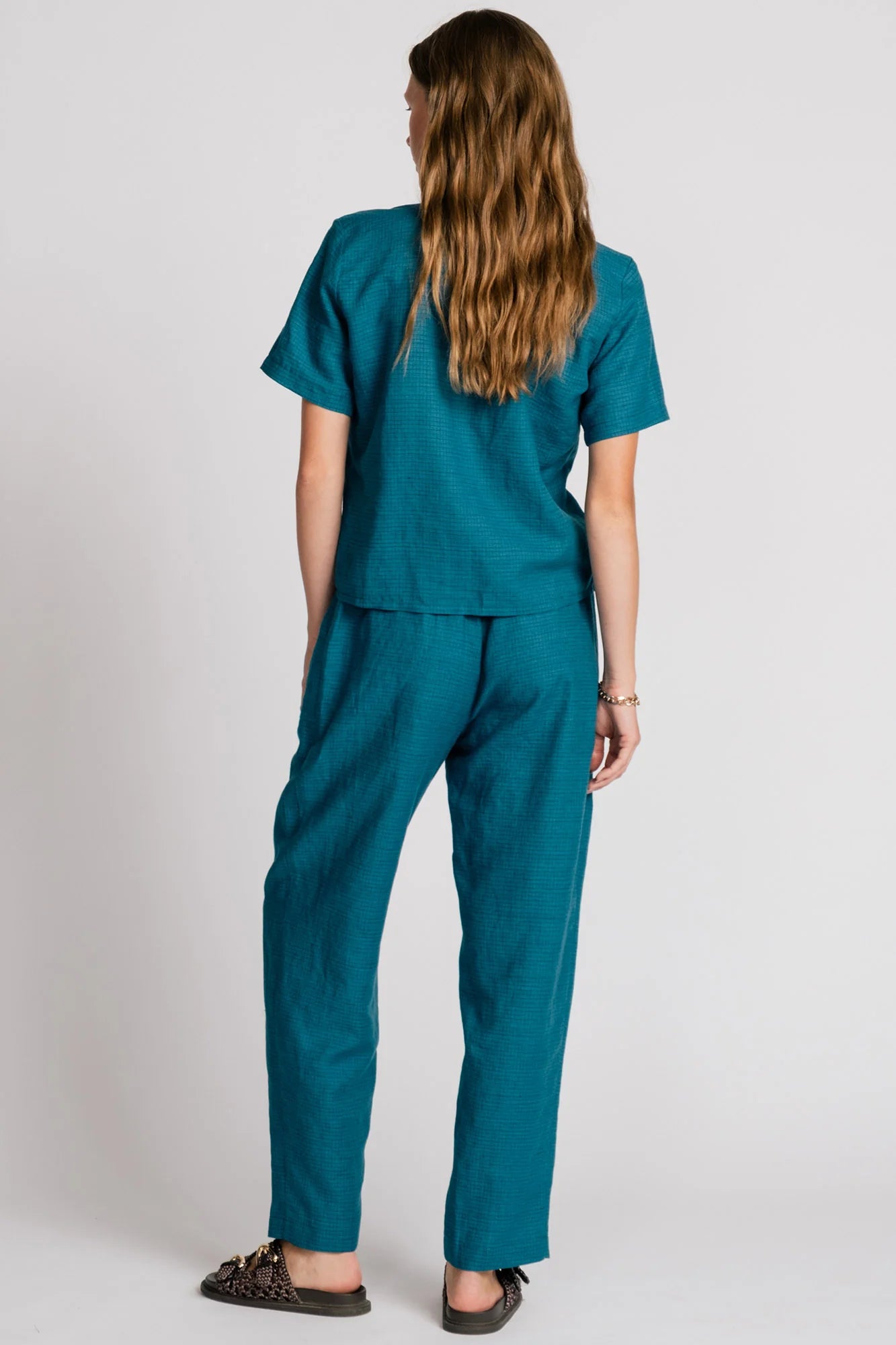 Back view of a woman wearing the Kite Blouse by Allison Wonderland in Teal with subtle check print, a blouse with a button front and a banded collar. It has  slightly loose fit and short sleeves. She is wearing it with matching pants and standing in front of a white background. 