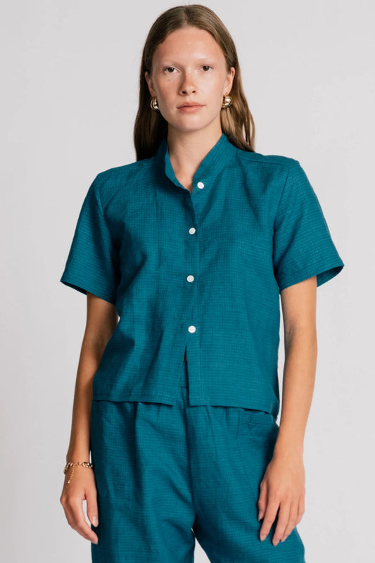 A woman wearing the Kite Blouse by Allison Wonderland in Teal with subtle check print, a blouse with a button front and a banded collar. It has  slightly loose fit and short sleeves. She is wearing it with matching pants and standing in front of a white background. 