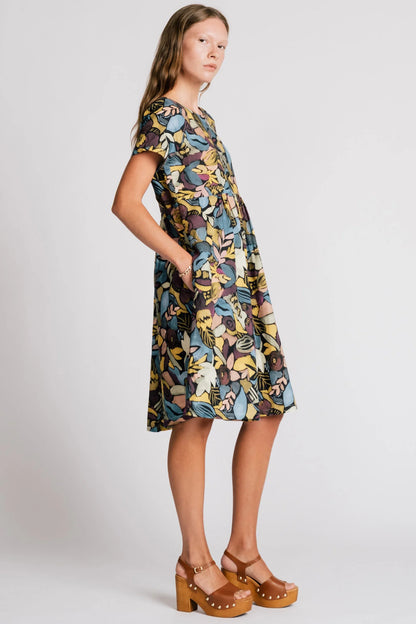 Side view of a woman wearing the Lark Dress by Allison Wonderland in Black Floral print, a short sleeved babydoll dress with pockets and a crossover back. She is standing in front of a plain background. 