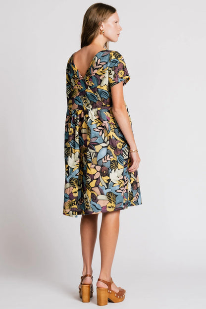 Back view of a woman wearing the Lark Dress by Allison Wonderland in Black Floral print, a short sleeved babydoll dress with pockets and a crossover back. She is standing in front of a plain background. 