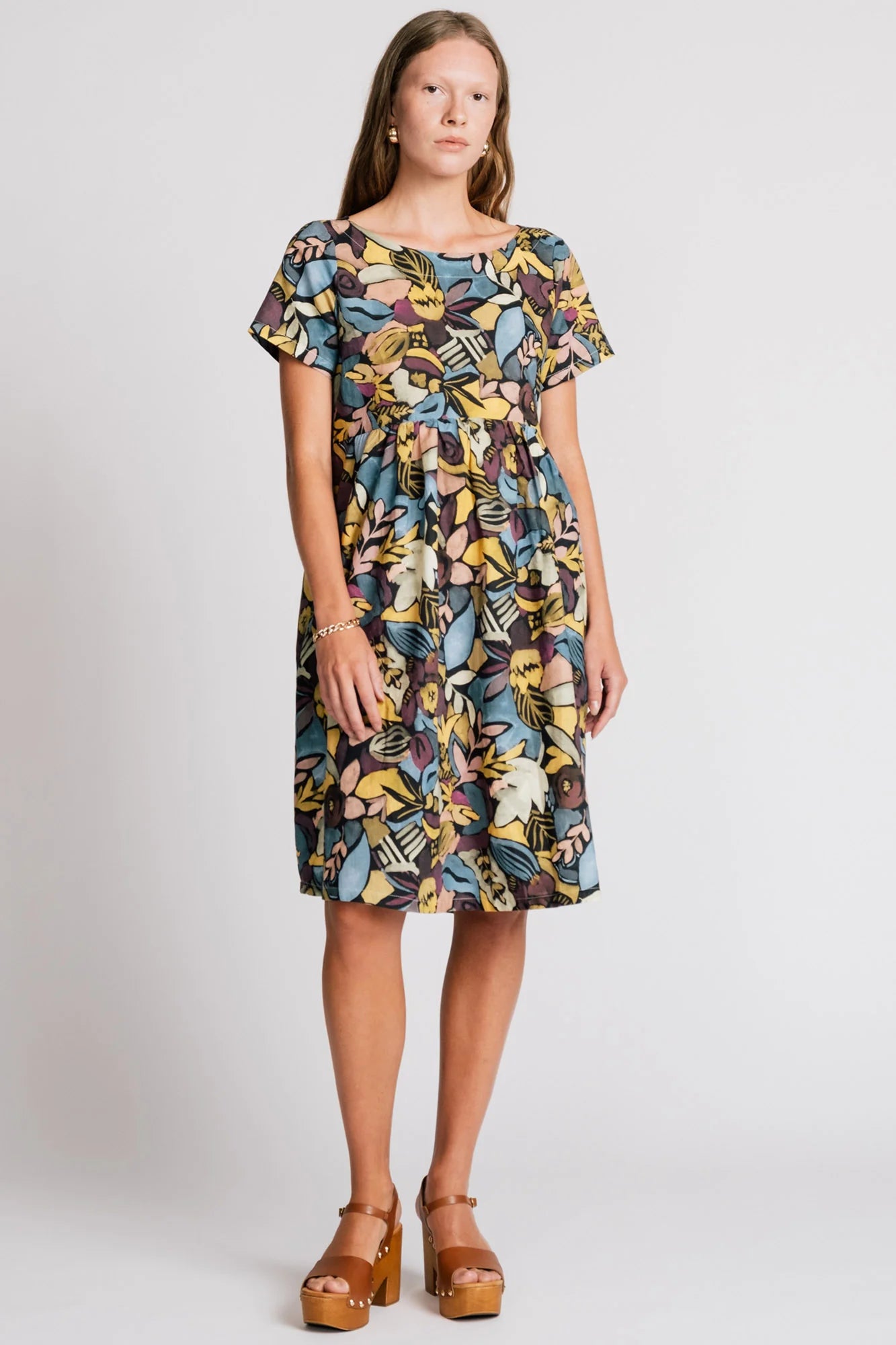 A woman wearing the Lark Dress by Allison Wonderland in Black Floral print, a short sleeved babydoll dress with pockets and a crossover back. She is standing in front of a plain background. 