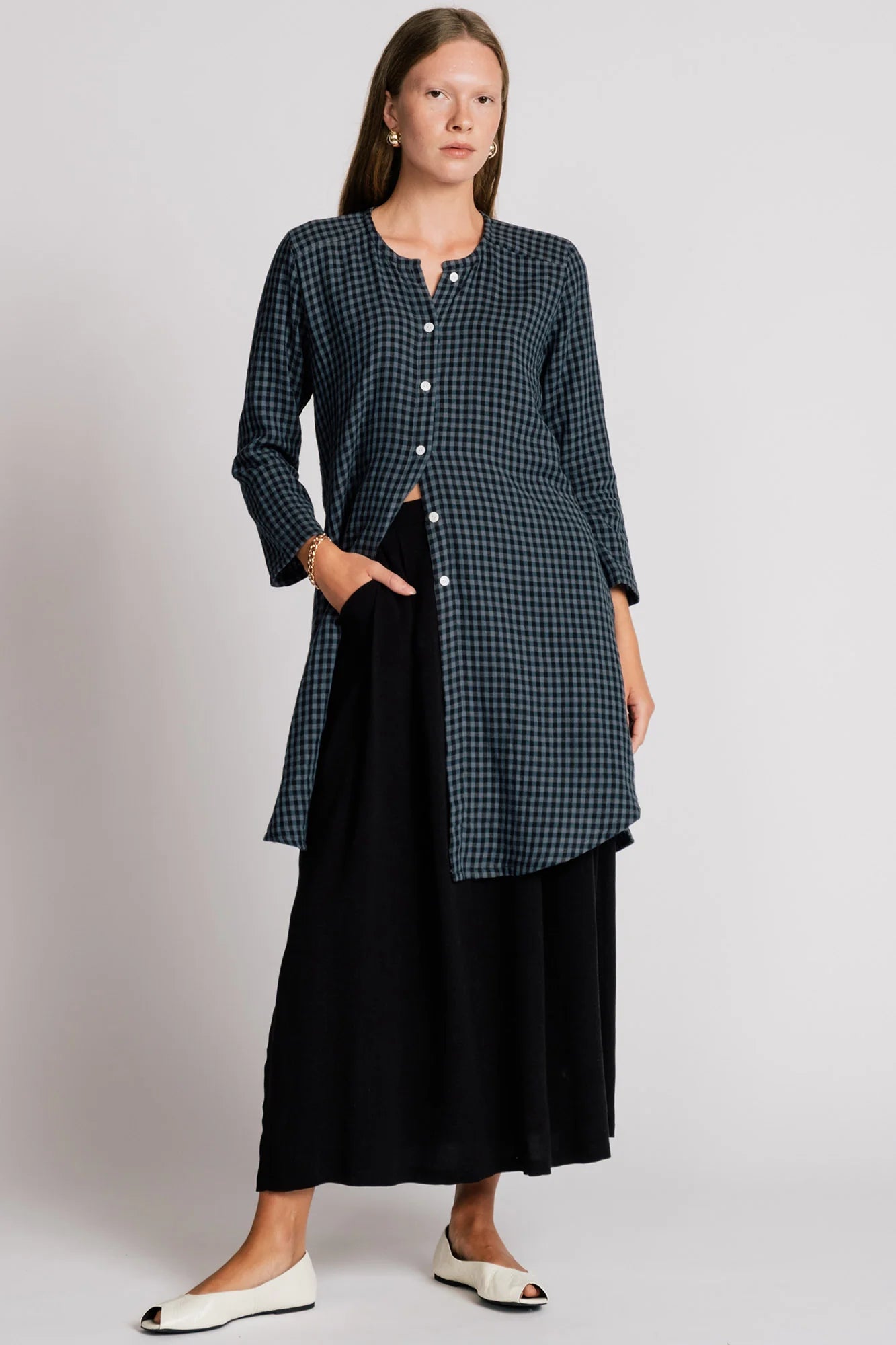 A woman wearing the Oriole Tunic by Allison Wonderland in Navy Gingham, a long tunic with a button front and rounded hi-low hemline. She is wearing it over a black skirt and standing against a grey background. 