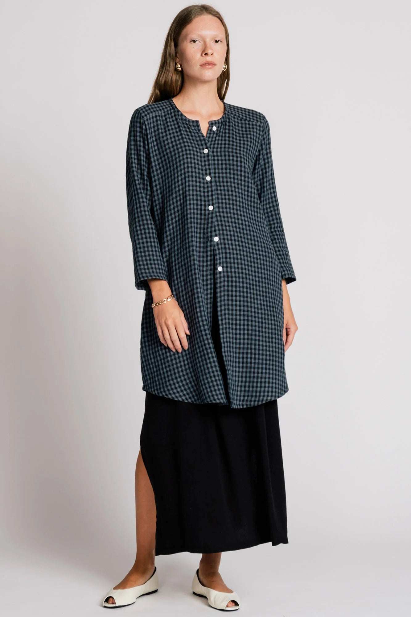 A woman wearing the Oriole Tunic by Allison Wonderland in Navy Gingham, a long tunic with a button front and rounded hi-low hemline. She is wearing it over a black skirt and standing against a grey background. 