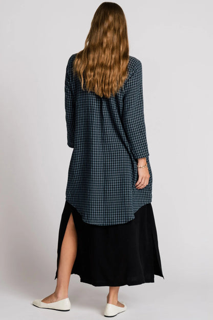 Back view of a woman wearing the Oriole Tunic by Allison Wonderland in Navy Gingham, a long tunic with a button front and rounded hi-low hemline. She is wearing it over a black skirt and standing against a grey background. 