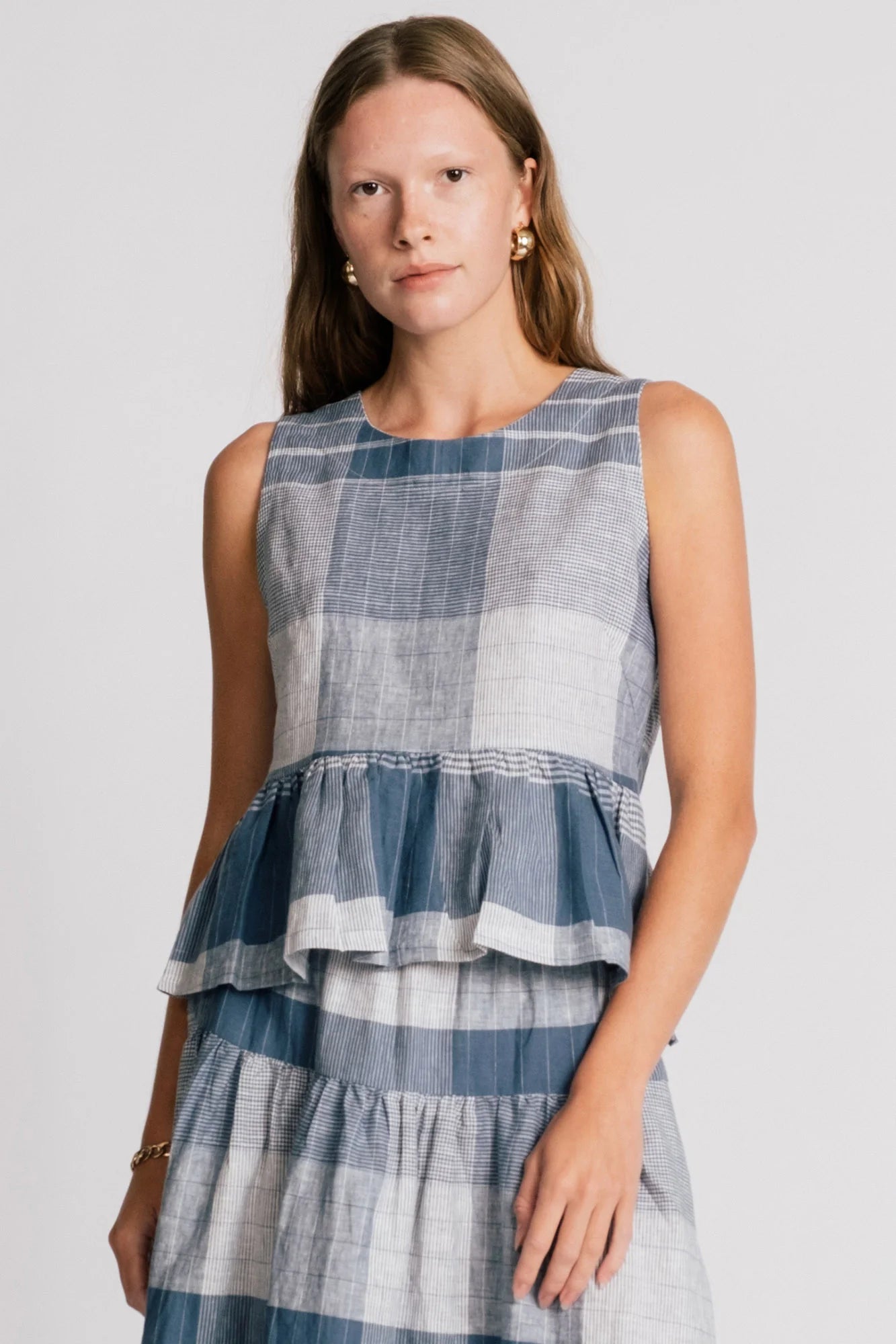 A woman wearing the Osprey Blouse by Allison Wonderland in Blue Plaid, a sleeveless top with a peplum both front and back and a cross-over back. She is wearing it with the matching Pelican Skirt and standing in front of a plain background.  