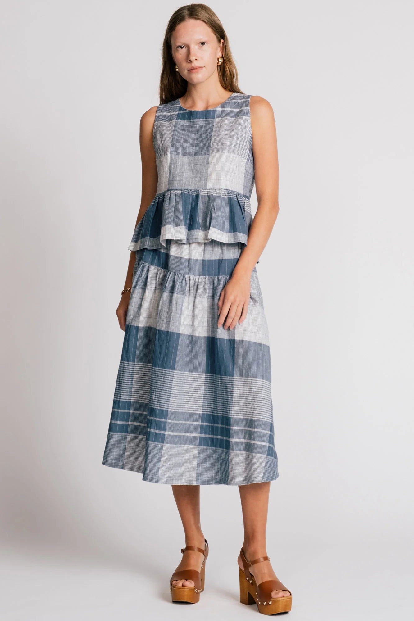 A woman wearing the Osprey Blouse by Allison Wonderland in Blue Plaid, a sleeveless top with a peplum both front and back and a cross-over back. She is wearing it with the matching Pelican Skirt and standing in front of a plain background. 