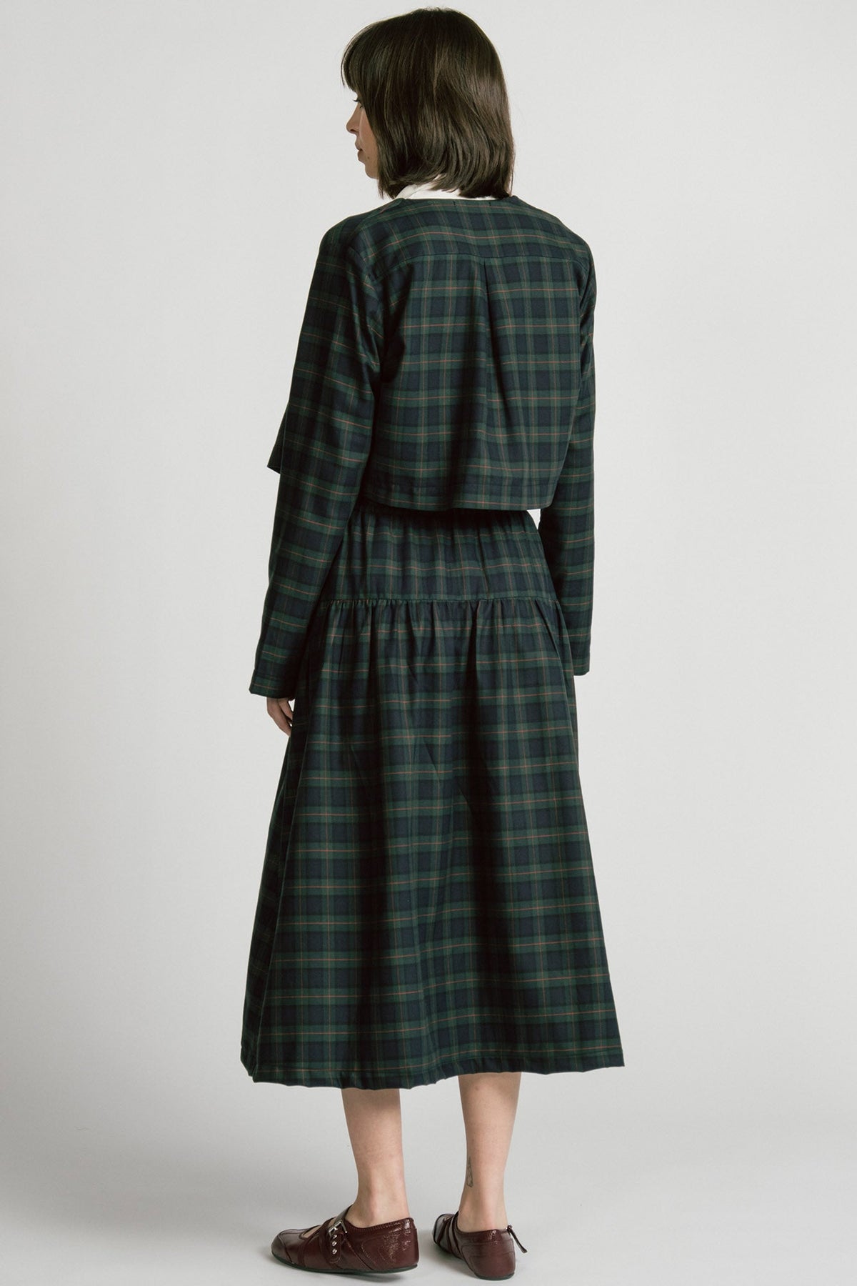Back view of a woman wearing the Pegasus Skirt in Plaid by Allison Wonderland, a midi length A-line skirt with an elastic waist, with the matching Libra bolero and a white top. She is standing in front of a white background