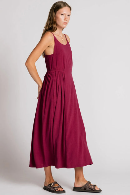 Side view of a woman wearing the Swan Dress by Allison Wonderland in Pink, a maxi dress with spaghetti straps, and ties at the waist. 