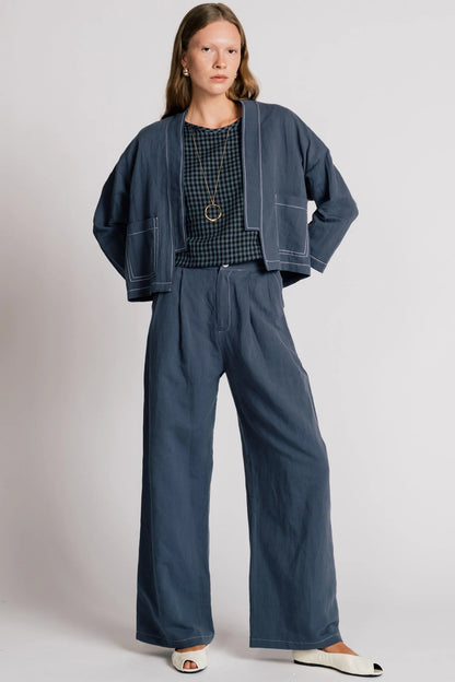 A woman wearing the Tern Blazer by Allison Wonderland in Navy, a cropped open jacket with a boxy fit and two front patch pockets. She is wearing it with the matching Whistler Pants and a gingham top and standing against a white background. 