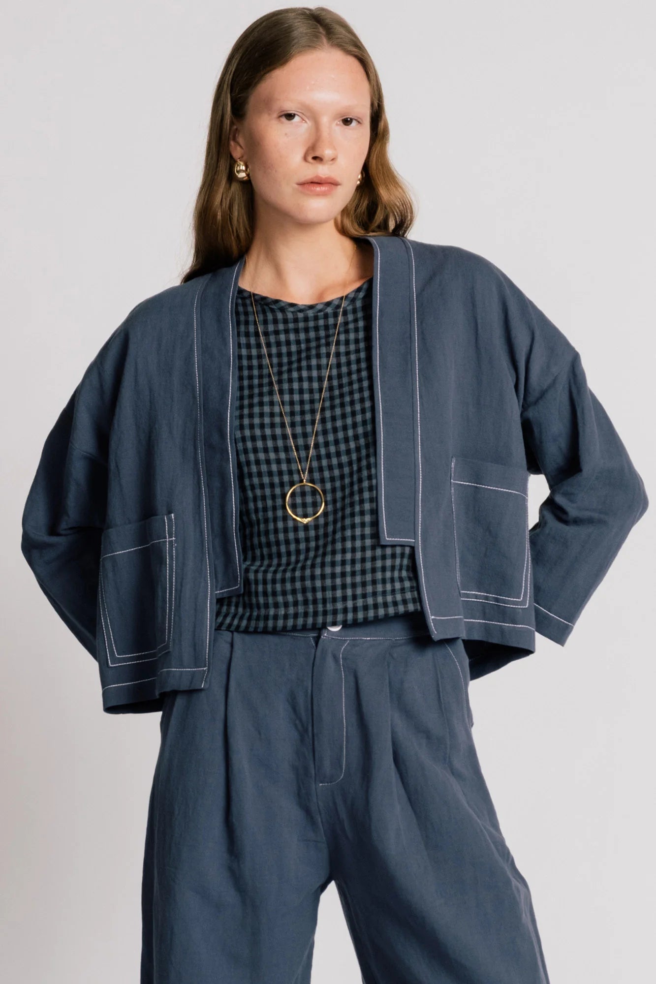 A woman wearing the Tern Blazer by Allison Wonderland in Navy, a cropped open jacket with a boxy fit and two front patch pockets. She is wearing it with the matching Whistler Pants and a gingham top and standing against a white background. 