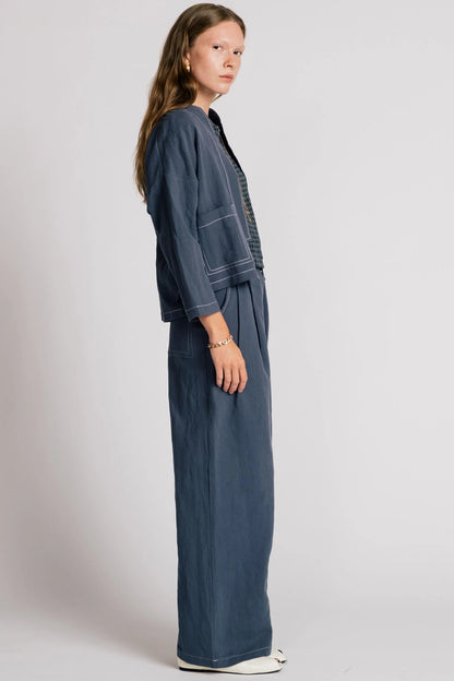 Side view of a woman wearing the Tern Blazer by Allison Wonderland in Navy, a cropped open jacket with a boxy fit and two front patch pockets. She is wearing it with the matching Whistler Pants and a gingham top and standing against a white background. 