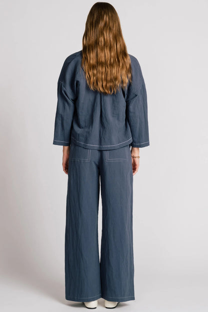 Back view of a woman wearing the Tern Blazer by Allison Wonderland in Navy, a cropped open jacket with a boxy fit and two front patch pockets. She is wearing it with the matching Whistler Pants and a gingham top and standing against a white background. 