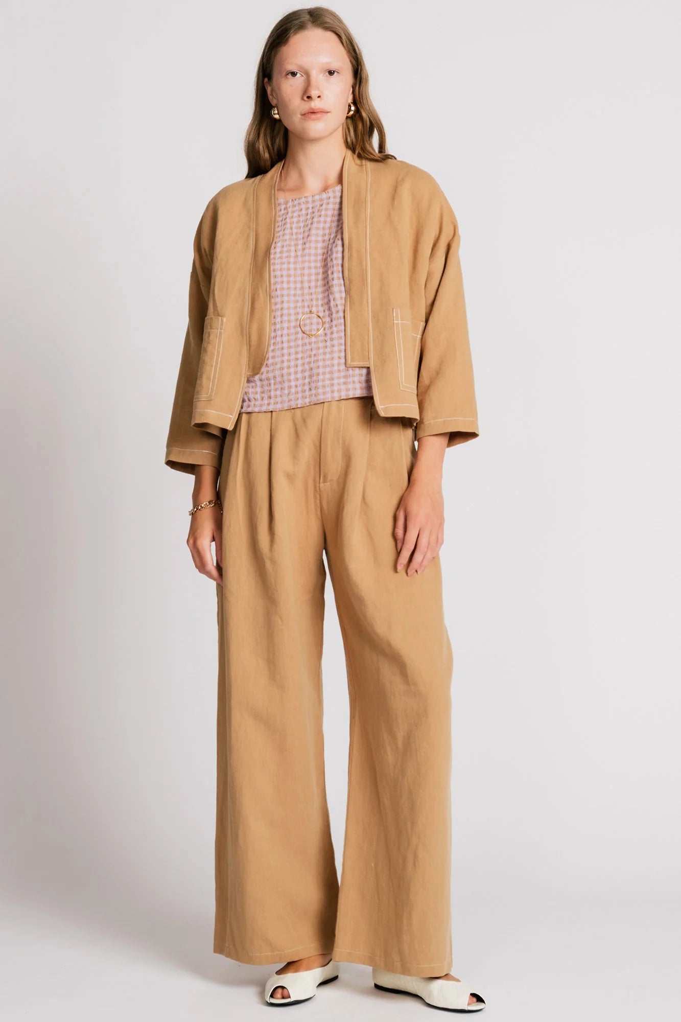 A woman wearing the Tern Blazer by Allison Wonderland in Sand, a cropped open jacket with a boxy fit and two front patch pockets. She is wearing it with the matching Whistler Pants and a light gingham top and standing against a white background. 