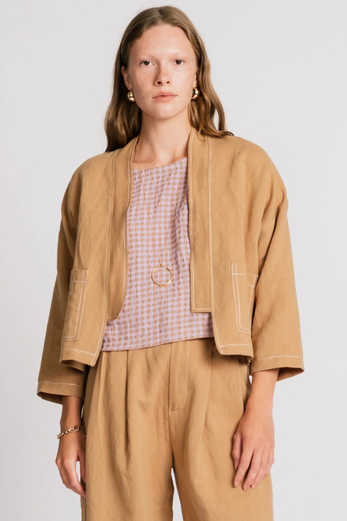 A woman wearing the Tern Blazer by Allison Wonderland in Sand, a cropped open jacket with a boxy fit and two front patch pockets. She is wearing it with the matching Whistler Pants and a light gingham top standing against a white background. 