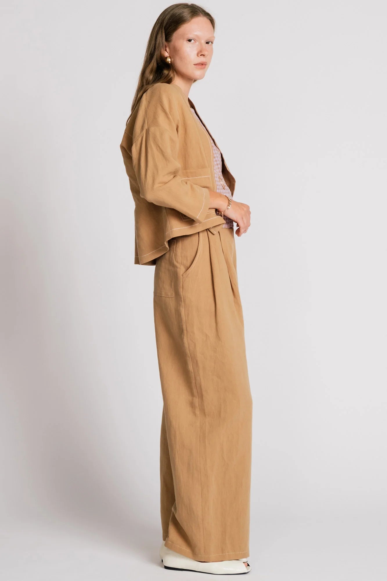 Side view of a woman wearing the Tern Blazer by Allison Wonderland in Sand, a cropped open jacket with a boxy fit and two front patch pockets. She is wearing it with the matching Whistler Pants and standing against a white background. 