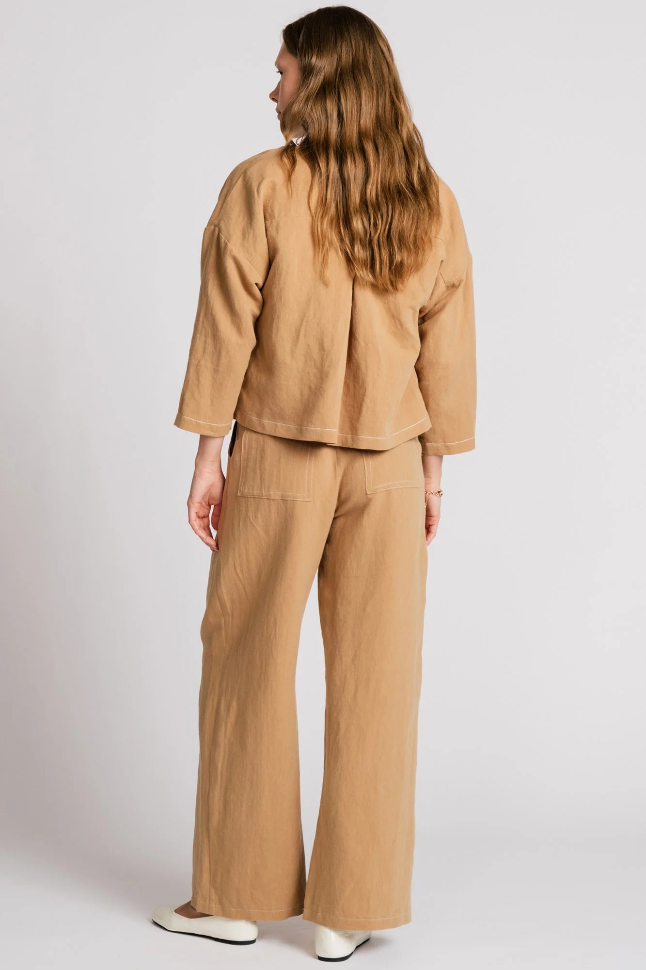 Back view of a woman wearing the Tern Blazer by Allison Wonderland in Sand, a cropped open jacket with a boxy fit and two front patch pockets. She is wearing it with the matching Whistler Pants and standing against a white background. 