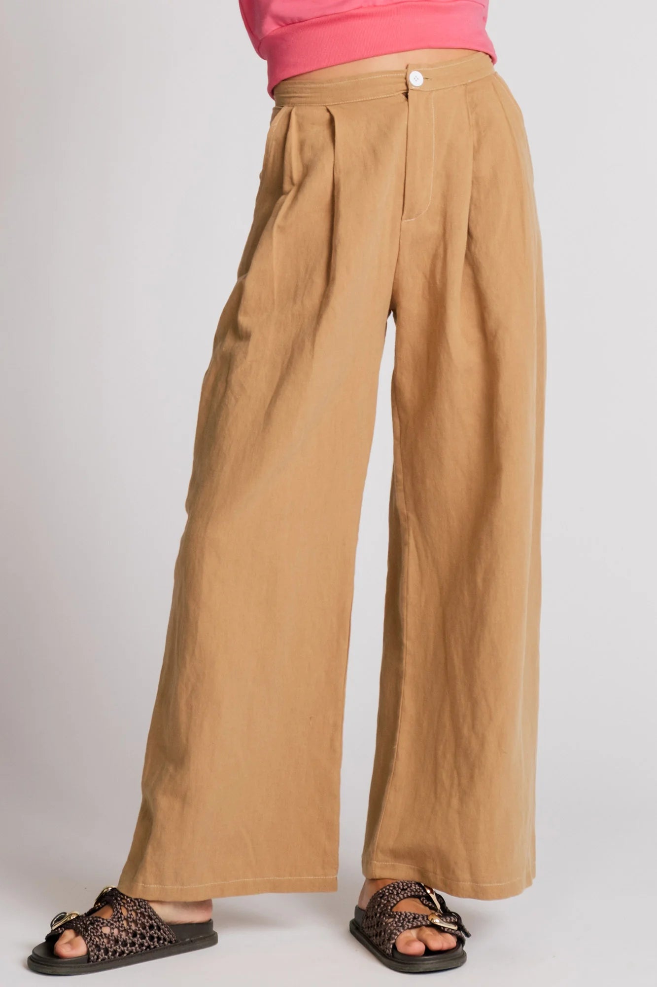  Waist down view of woman wearing the Whistler Pants by Allison Wonderland in Sand, wide legged pants with front pleats, a zip fly, and contrast white stitching. She is wearing them with a pink top and standing in front of a white background. 