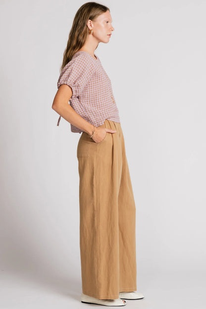 Side view of a woman wearing the Whistler Pants by Allison Wonderland in Sand, wide legged pants with front pleats, a zip fly, and contrast white stitching. She is wearing them with a light gingham top and standing in front of a white background. 