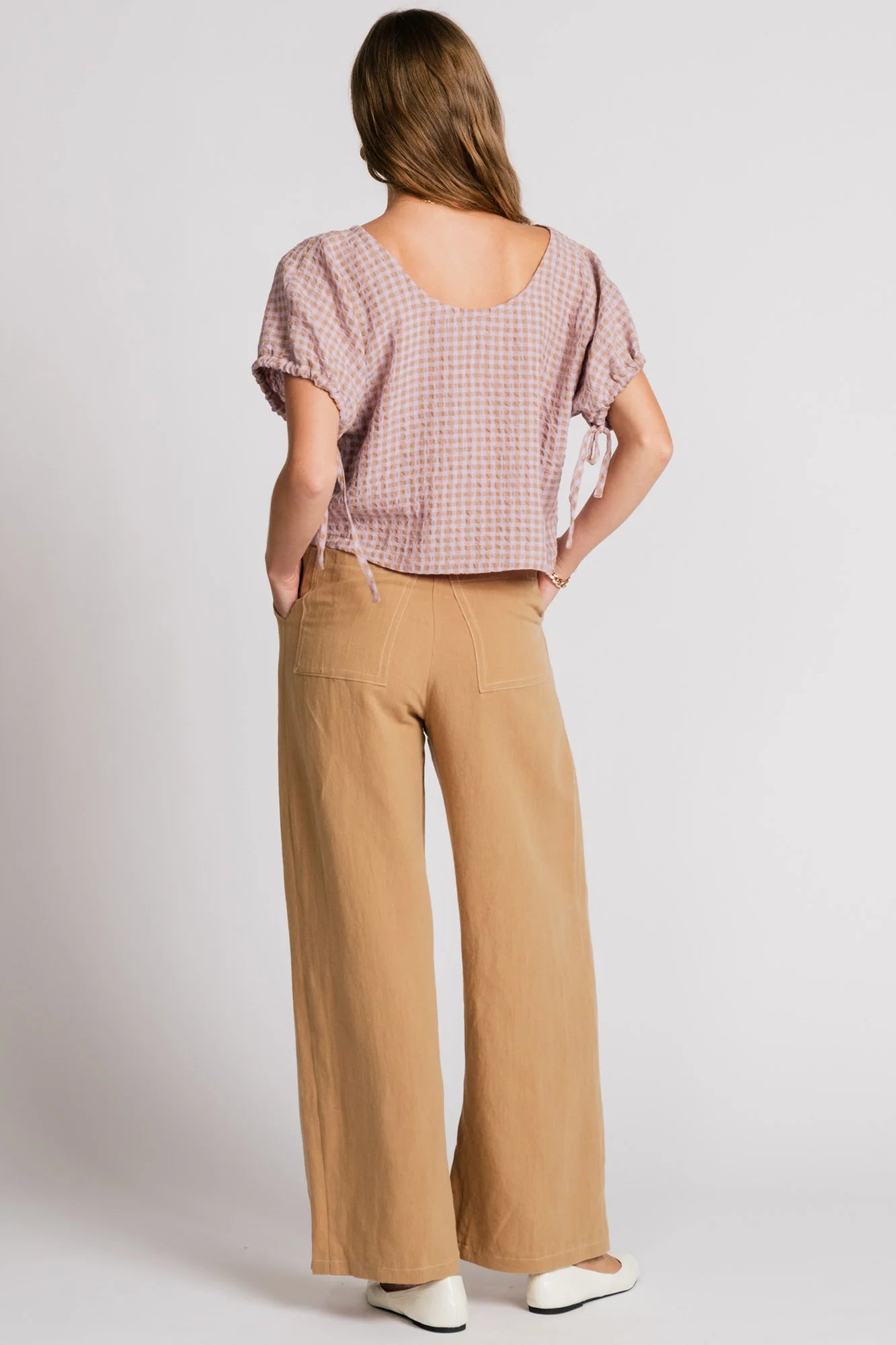 Back view of a woman wearing the Whistler Pants by Allison Wonderland in Sand, wide legged pants with front pleats, a zip fly, and contrast white stitching. She is wearing them with a light gingham top and standing in front of a white background. 