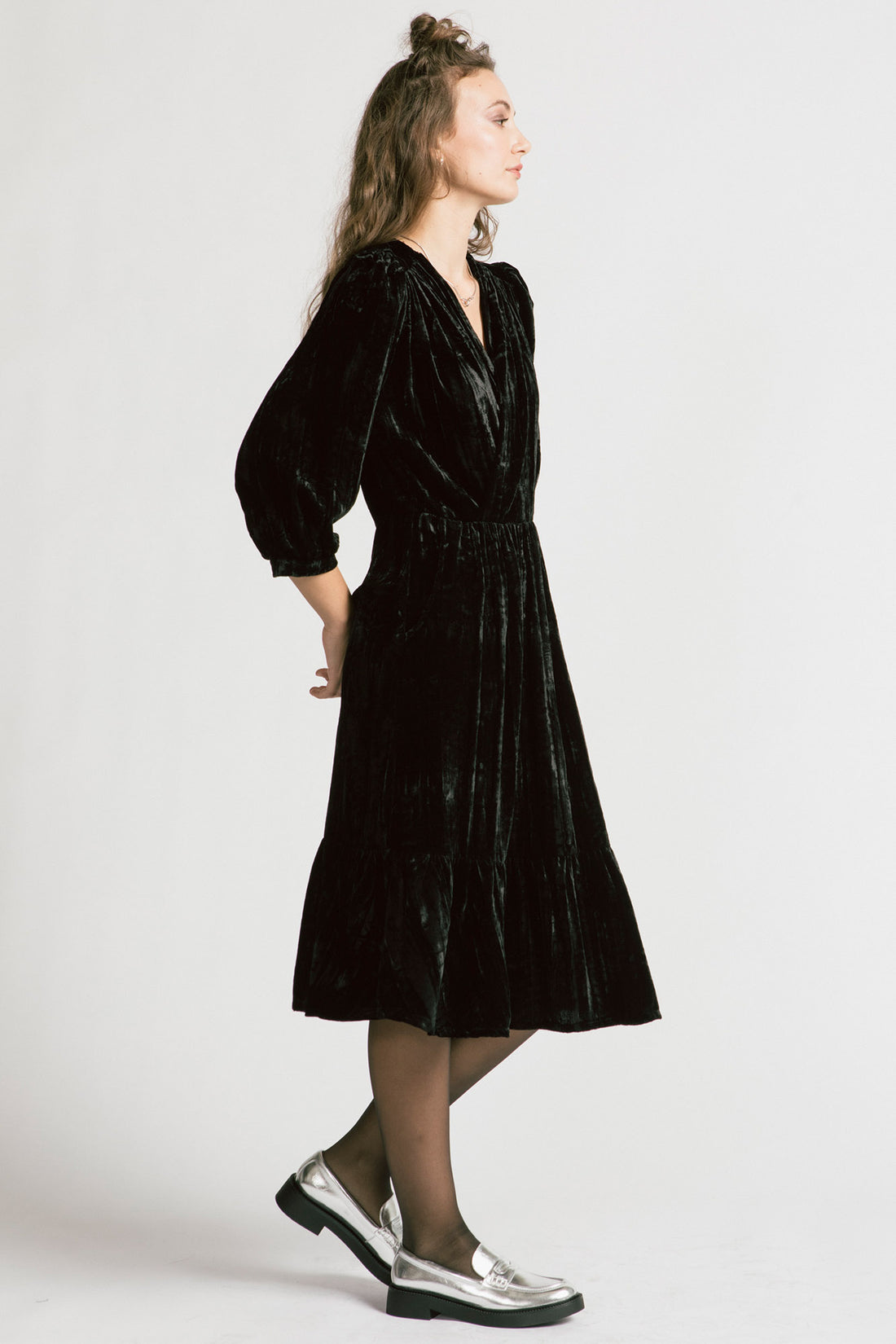 Side view of a woman wearing the Dark Lover Dress by Allison Wonderland in Black crushed velvet, featuring a faux-wrap front, 3/4 gathered sleeves, and a tiered midi-length skirt. She is standing in front of a white background. 