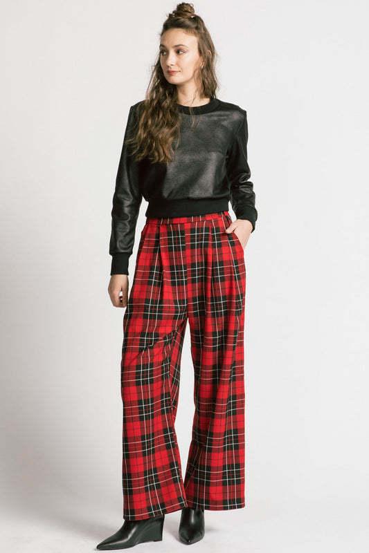 A woman wearing the Gone Top by Allison Wonderland in Leatherette, a classic crewneck in faux leather fabric, with red plaid pants. She is standing in front of a white background 