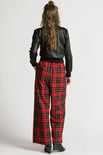 Back view of a woman wearing the Gone Top by Allison Wonderland in Leatherette, a classic crewneck in faux leather fabric, with red plaid pants. She is standing in front of a white background 