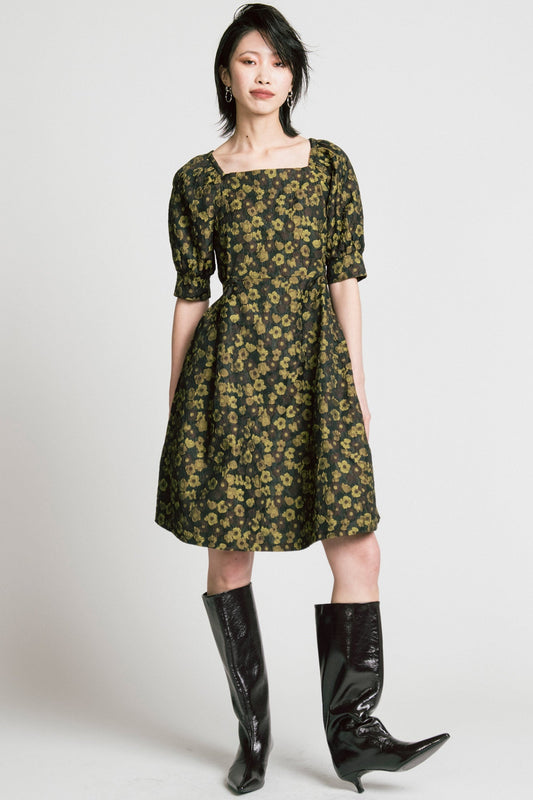 A woman wearing the Priority Dress by Allison Wonderland in Floral Jacquard Knit, with a square neck, a baby doll shape, side ties, elbow-length sleeves, and a below the knee hem. She is standing in front of a grey background. 