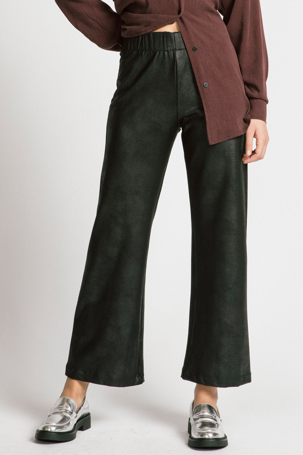 Waist down view of a woman wearing the Shanna Pants by Allison Wonderland in Black Leatherette, loose fitting flared pants with an elastic waistband. She is wearing them with a brown top and standing in front of a silver background. 