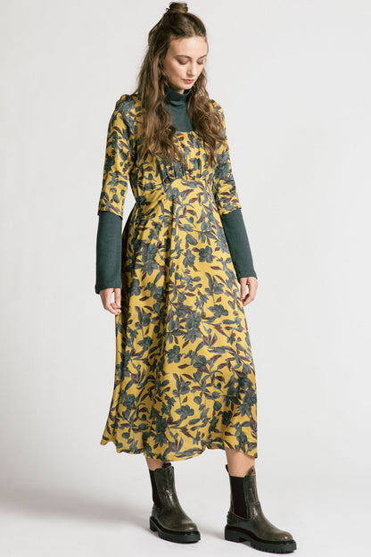 A woman wearing the Tempest Dress by Allison Wonderland in Gold Floral, with a  scoop neck, cinched empire waist, 3/4 sleeves, and long full skirt. She's wearing it over the Forever Turtleneck and is standing in front of a white background. 
