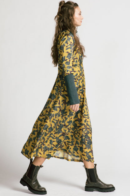 Side view of a woman wearing the Tempest Dress by Allison Wonderland in Gold Floral, with a  scoop neck, cinched empire waist, 3/4 sleeves, and long full skirt. She's wearing it over the Forever Turtleneck and is standing in front of a white background. 