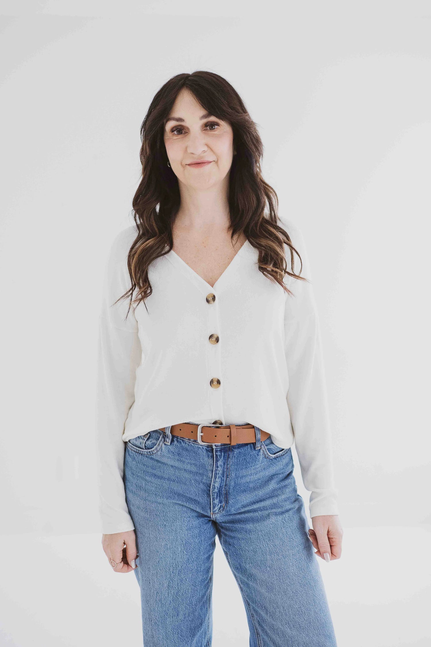 A woman wearing the Holly Cropped Cardigan by Ang Hill in Vanilla, a v-neck cardigan with drop sleeves and tortoiseshell buttons. She is wearing it with jeans and is standing in front of a white background. 