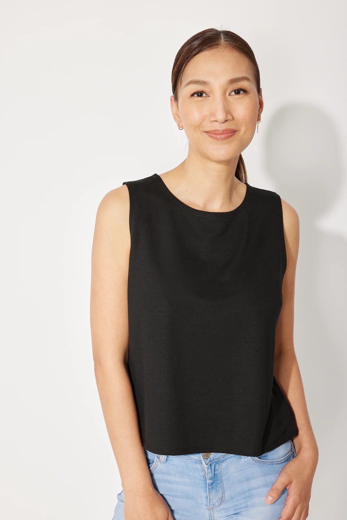 A woman wearing the Baie Cami by Canopée in Black, a bamboo French Terry tank with a high neck and loose fit. She is wearing it with jeans and standing in front of a white background. 