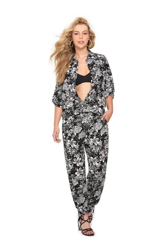 A woman wearing the Batan Pants by Luc Fontaine in  Black and White Tropical Print, featuring an elastic waist, relaxed fit, and ties at the ankles She is wearing them with the matching Babuyan Blouse and standing in front of a white background. 