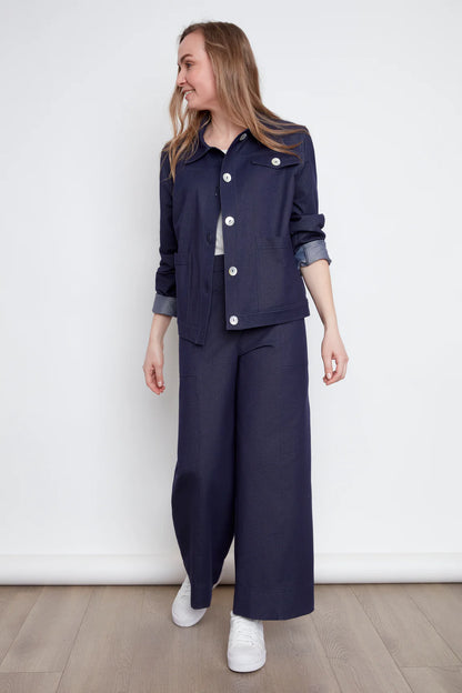 A woman wearing the Bella Knit Jacket by Julei, a denim style jacket with large contrast buttons, a chest pocket and two front patch pockets. She is wearing it with a white and the matching Brooke Knit Trousers and standing in front of a white wall. 