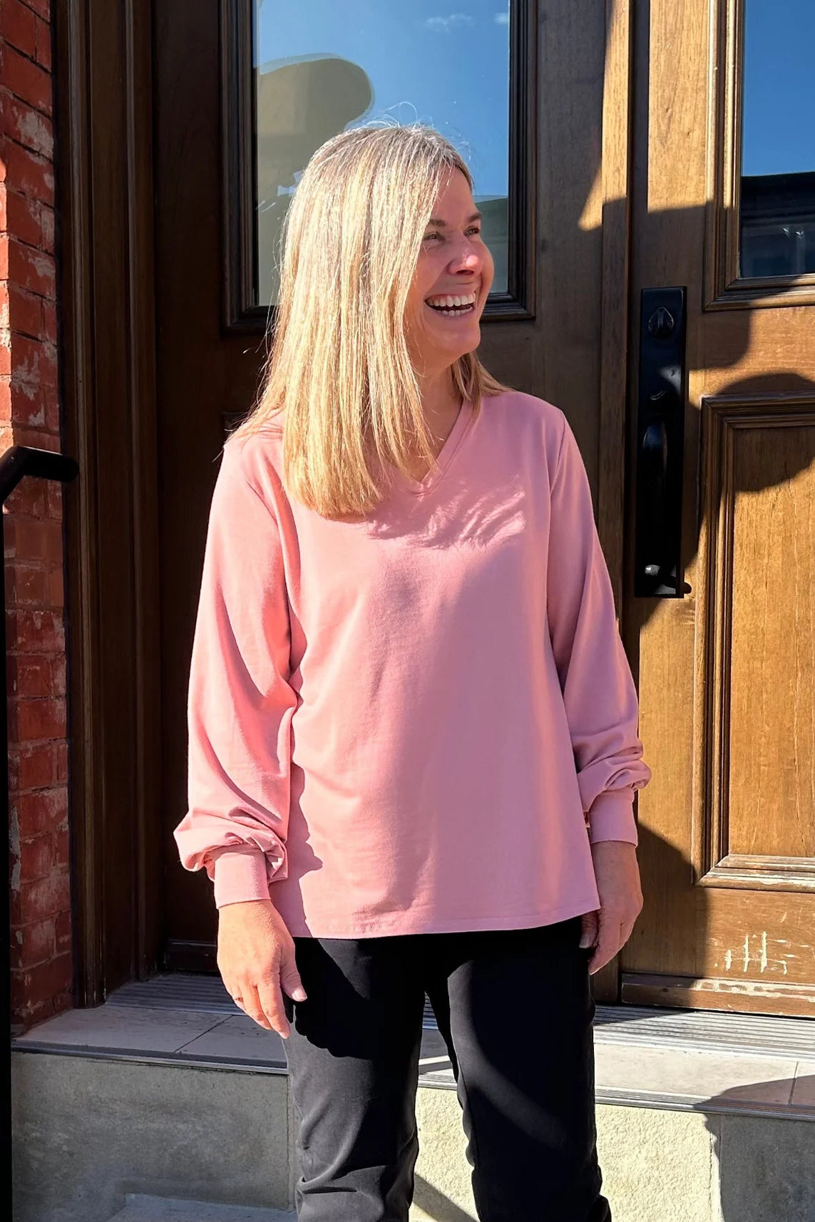 A woman wearing the Belmina Long Sleeve Top by Message Factory in Pink, with a small v-neck, long sleeves with puffed cuffs, and a relaxed fit. She is wearing it with black pants and standing outside in front of a wooden door. 