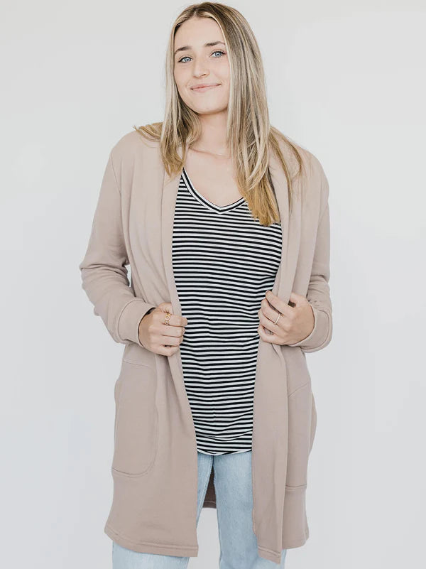 A woman wearing the Blondie Classic Briton Cardigan in Stucco, a long open front cardigan with large patch pockets. She is wearing it with jeans and a striped t-shirt and standing in front of a white background. 