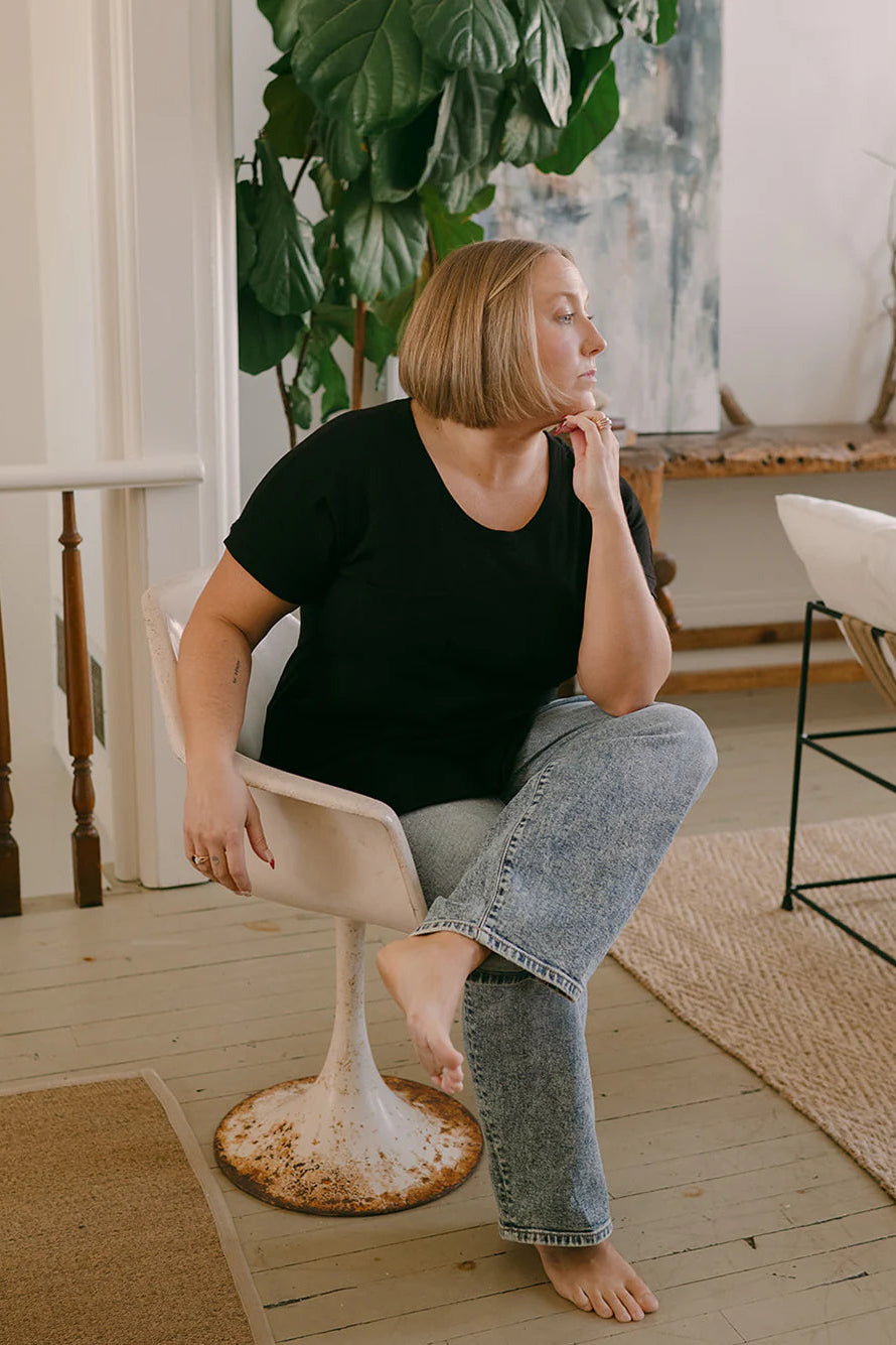 A woman wearing the Janis Tee by Blondie in Black Slub, with a round neck, short sleeves, breast pocket, and hi-low hemline. She is wearing it with jeans and sitting in a chair. 