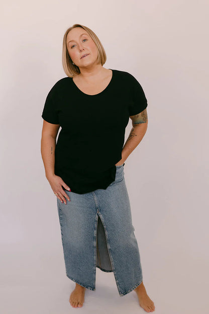 A woman wearing the Janis Tee by Blondie in Black Slub, with a round neck, short sleeves, breast pocket, and hi-low hemline. She is wearing it with a jean skirt and standing in front of a white background. 